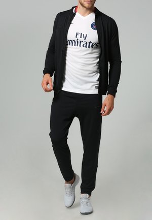Hombre con chaqueta negra con cremallera, camiseta blanca del Paris Saint-Germain, pantalones negros y zapatillas blancas, de pie contra un fondo gris.