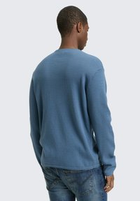 Blaues Strickpullover mit strukturiertem Rippmuster und langen Ärmeln, getragen mit blauen Jeans, von hinten gesehen.