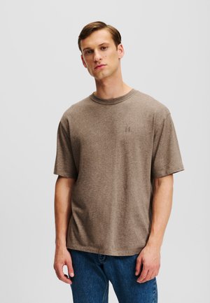 Tricou din bumbac maro, cu mâneci scurte, decolteu rotund și detaliu cusut cu logo pe piept, purtat cu blugi albaștri.