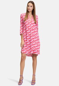 Gemustertes Kleid in lebhaften Rosa- und Weißtönen, mit lockerer Passform, drei Viertelarm und V-Ausschnitt mit orangefarbener Einfassung, kombiniert mit lila Absätzen.