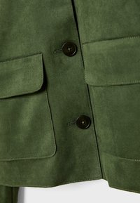 Giacca di camoscio verde con due tasche a pattina, una texture liscia e due bottoni neri per la chiusura. I dettagli delle cuciture sono visibili.