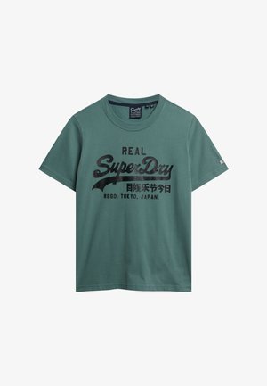 Zielona koszulka z krótkim rękawem i okrągłym dekoltem, z czarnym logo "Real SuperDry Regd. Tokyo, Japan" oraz japońskimi znakami na klatce piersiowej.