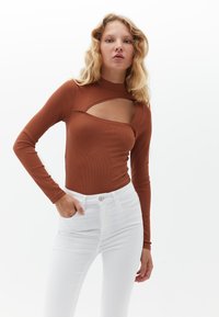 Braunes, geripptes Langarmoberteil mit einem Ausschnitt-Design, kombiniert mit weißen, hochgeschnittenen Jeans mit Reißverschluss und Knopfverschluss.