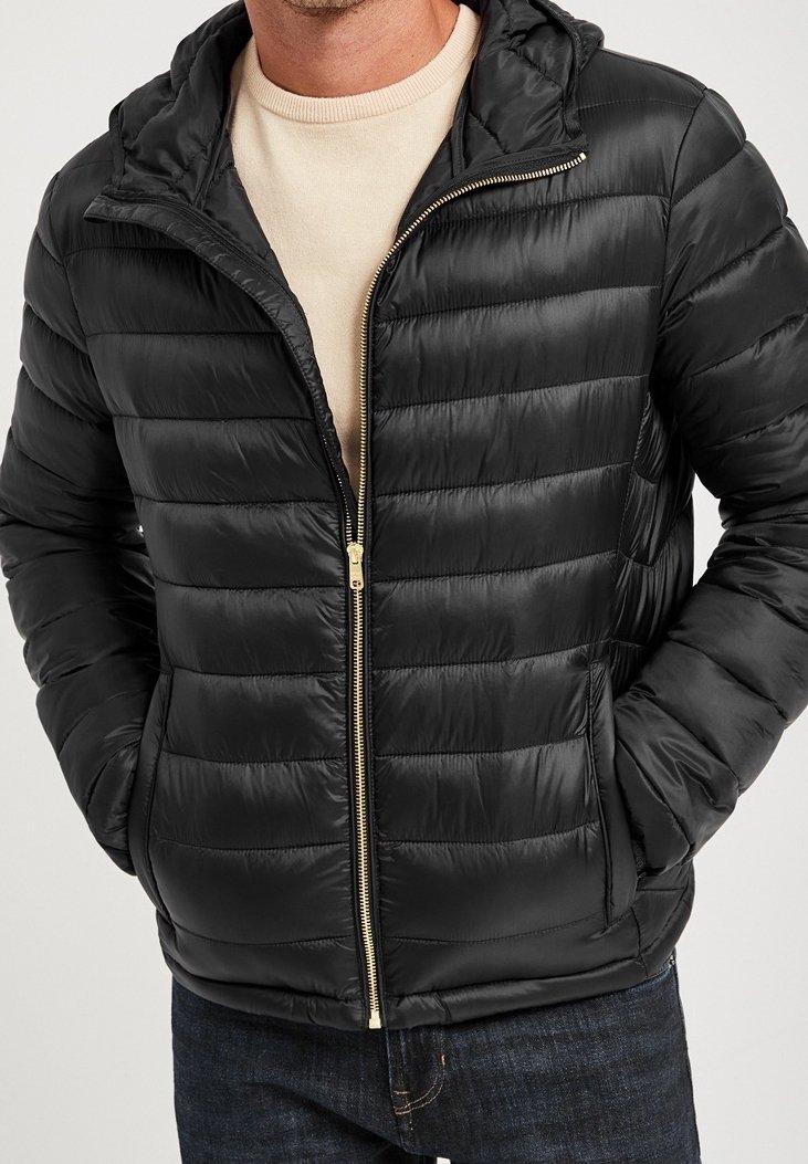 Man i en svart vadderad jacka med dragkedja över en ljus tröja, med händerna i fickorna, mot en neutral bakgrund.