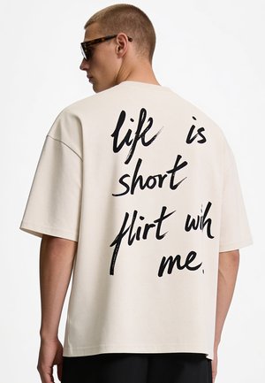 Jeune homme portant des lunettes de soleil et un t-shirt blanc oversize avec un texte manuscrit noir disant "life is short flirt with me" dans le dos.