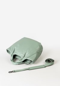 Borsa a mano in pelle verde chiaro con una superficie morbida, forma rettangolare e manico arrotolato. Tracolla regolabile posata accanto alla borsa.