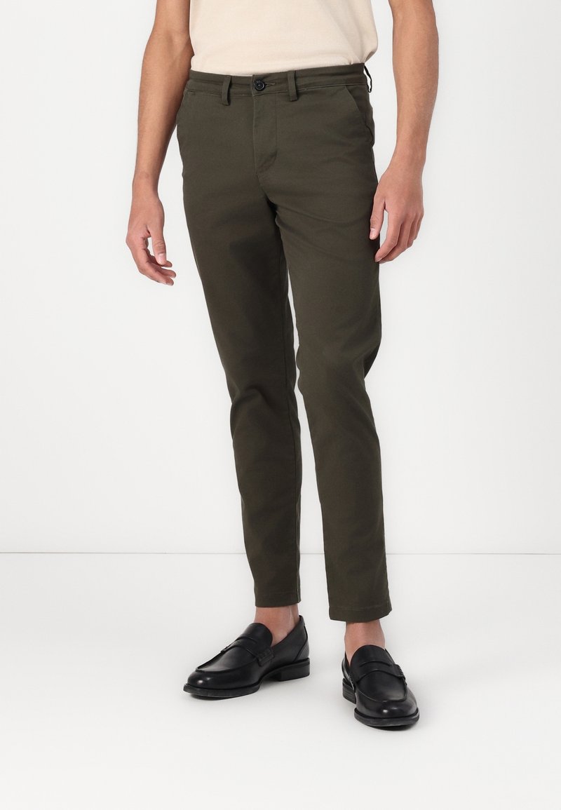 Pantalons chinos verts olive à coupe slim, en coton, avec une fermeture à boutons, des passants pour ceinture et des poches latérales, associés à des mocassins noirs.