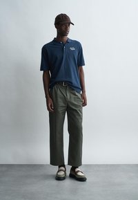 Hombre con camiseta polo navy, pantalones cortos de color verde oliva, gorra marrón clara, calcetines blancos y mocasines beige, de pie contra un fondo gris liso.
