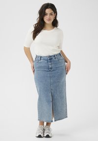 Falda maxi de mezclilla en azul claro con abertura frontal, cintura alta y cinco bolsillos. Combinada con una blusa blanca y zapatillas grises.