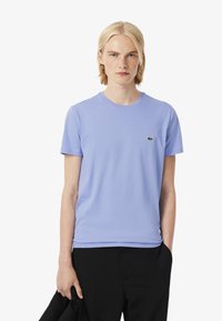 Een model draagt een lichtpaarse t-shirt met een klein groen logo, terwijl ze in de ene hand een zwarte jas vasthoudt en tegen een effen witte achtergrond staat.