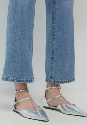 Piedi che indossano scarpe basse argentate a punta con cinturini alla caviglia e jeans blu a gamba larga su uno sfondo semplice.