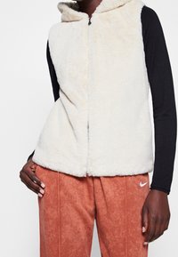 Béžový kožešinový vest z umělé kožešiny s kapucí, který má přední zip. Kombinovaný s oranžovo-hnědými strukturovanými kalhotami a černým topem s dlouhým rukávem.