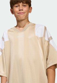 Chlapec s krátkymi hnedými vlasmi, oblečený v béžovom športovom tričku Adidas s bielymi a svetlohnedými panelmi na rukávoch.