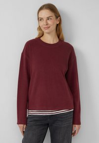 Vrouw met licht haar, gekleed in een donkerrode trui met lange mouwen over een gestreept shirt en donkere jeans, staande tegen een effen lichtmuur.