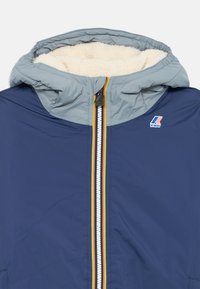 Veste bleu marine avec une capuche doublée en polaire grise, dotée d'un zip central avec une bande colorée et d'un logo K-Way sur la poitrine.