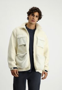 Beżowa polarowa kurtka z zamkiem z przodu, dwiema kieszeniami na piersi i dwiema kieszeniami dolnymi. Posiada wysoki kołnierz i kontrastowe akcenty z tkaniny.