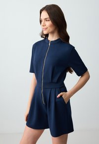 Navyblauwe korte romper van soepel materiaal, met een half-rits voorkant, korte mouwen en een getailleerde taille met zijzakken.