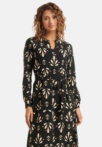 Schwarzes Langarmkleid mit V-Ausschnitt und Bindegürtel, das mit einem cremefarbenen Blumenmuster und roten Akzenten im gesamten Stoff versehen ist.