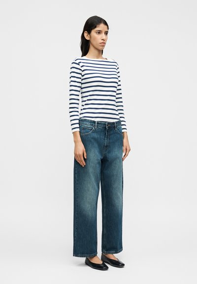 Ριγωτό πουκάμισο μακρυμάνικο σε λευκό και σκούρο μπλε, σε συνδυασμό με φαρδιά, ξεβαμμένα μπλε τζιν παντελόνια και μαύρα επίπεδα παπούτσια.