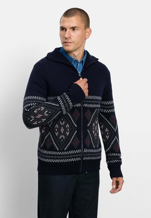 Uomo che indossa un maglione blu scuro con zip e motivi geometrici sopra una camicia blu con bottoni, in piedi contro uno sfondo chiaro e uniforme.