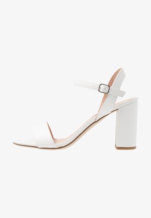 Sandalia de tacón blanco con tacón cuadrado, correa ajustable en el tobillo y banda frontal ancha, fabricada con un material sintético suave y plantilla neutra.