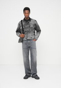 Juoda ir pilka džinsinė striukė su priekinėmis kišenėmis, derinama su pilkomis platėjusiomis džinsinėmis kelnėmis. Modelis laiko mažą juodą krepšį per petį.