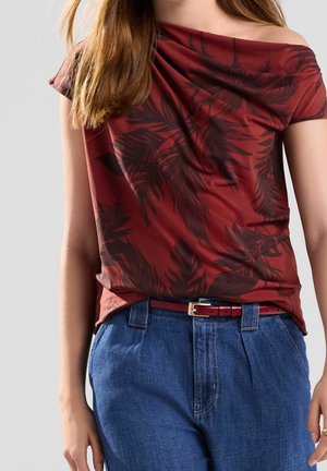 Jeune personne portant un haut bordeaux à épaules dénudées avec des motifs de feuilles noires et un pantalon en jean bleu taille haute avec une ceinture bordeaux.