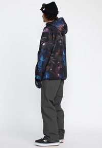 Veste de snowboard imperméable noire avec un motif floral coloré, associée à un pantalon de neige gris et des sneakers noires avec des accents blancs.