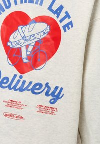 Lichtgrijze sweatshirt met blauwe tekst, een rood hart en een afbeelding van een sandwich op een fiets. Inclusief winkelinformatie in kleine rode letters.
