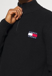 Suéter negro de punto con un cuello alto, que cuenta con una media cremallera y un parche de logo en azul, blanco y rojo en el pecho. Tejido texturizado.