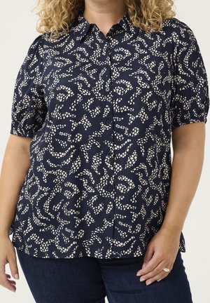 Kvinde iført en mørkeblå bluse med knapper og et abstrakt hvidt prikket mønster samt korte pufærmer, kombineret med mørkeblå jeans.