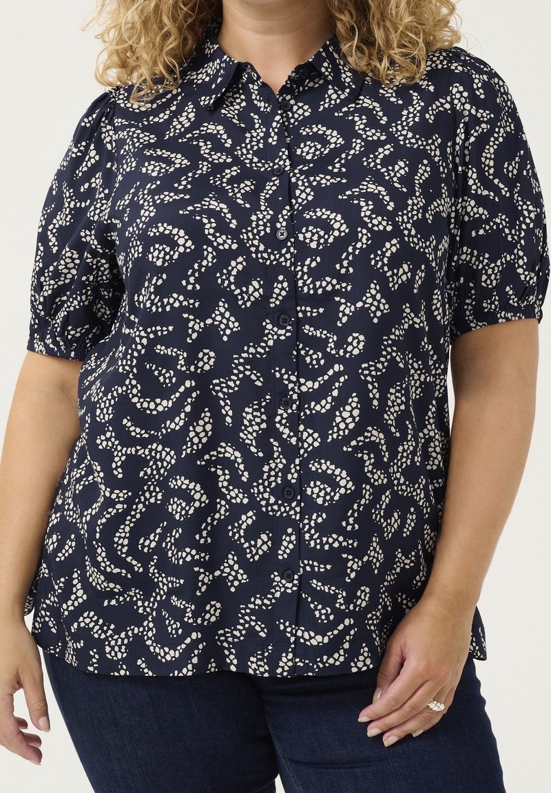 Femme portant un chemisier bleu marine à boutons avec un motif abstrait de points blancs et des manches courtes bouffantes, associé à un jean bleu foncé.