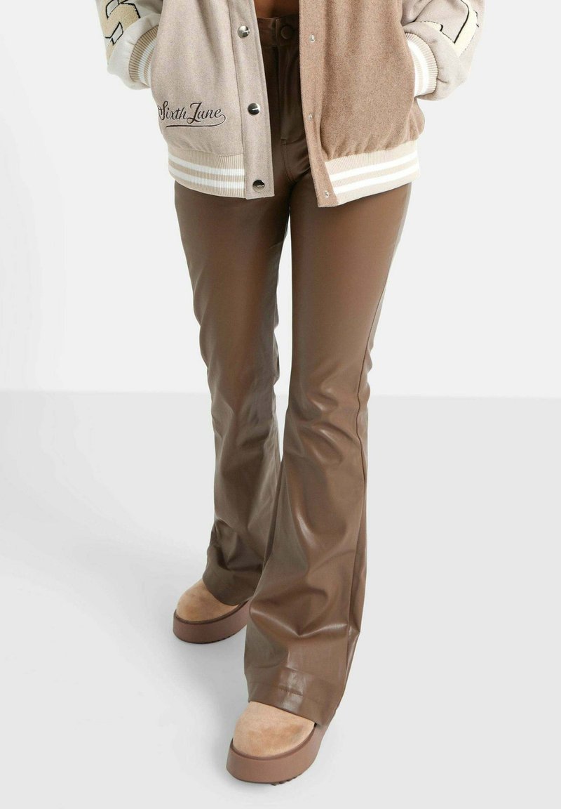 Bruine faux leren flarebroeken met een glanzende afwerking, gecombineerd met beige plateau-schoenen en een bicolor bomberjack in grijs en tan.