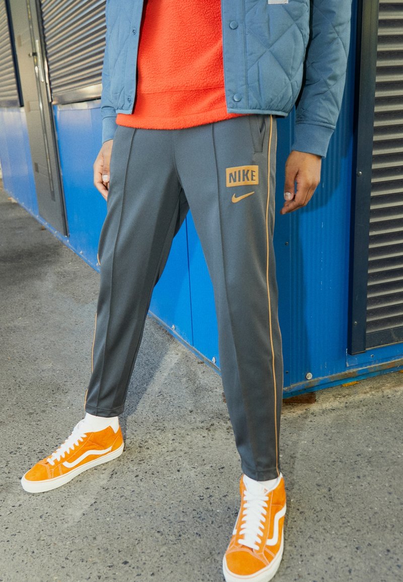 Grå atletiska byxor med orange Nike-logotyp och detaljer, kombinerat med orange mockaskor, bärs med en röd fleece-tröja och en blå jacka.