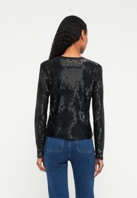 Femeie cu păr lung și ondulat, purtând un top lung cu textură neagră și blugi albaștri, văzută din spate pe un fundal simplu.