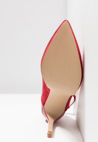 Chaussure à talon haut en daim rouge avec bout pointu et semelle beige lisse. Dispose d'un talon stilettos fin et d'une finition texturée.