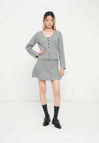 Szary wełniany kardigan z długimi rękawami i pięcioma guzikami, w połączeniu z szarą spódnicą w kształcie litery A. Modelka ma na sobie czarne buty i ciemnoszare skarpetki.