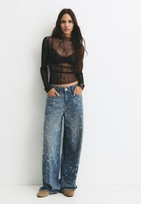 Top lung cu mânecă lungă, transparent, de culoare neagră, cu un model floral peste un sutien negru, asortat cu blugi largi, deschiși la culoare, cu un finisaj uzat.