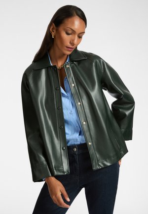Giacca in ecopelle verde con colletto ampio, bottoni a pressione e vestibilità rilassata, abbinata a una camicia a righe azzurre leggere.