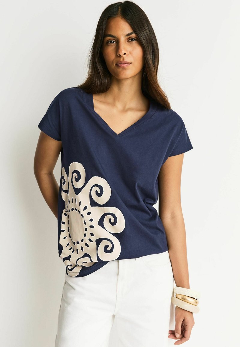 Granatowy bawełniany T-shirt z dekoltem w serek i krótkimi rękawami, z dużym kremowym wzorem wirującego słońca na przodzie. Białe spodnie i bransoletki.