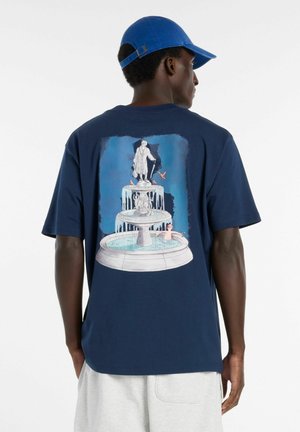 Mann mit blauer Kappe trägt ein marineblaues T-Shirt mit einer Illustration eines dreistufigen Brunnens mit Statuen und fließendem Wasser.