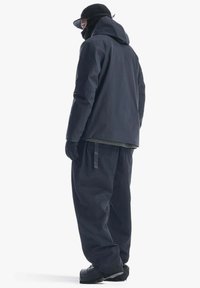 Giacca nera impermeabile con cappuccio, pantaloni larghi abbinati, guanti e berretto nero. Presenta un design minimalista e una finitura texturizzata.