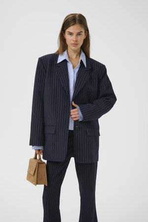 Navy pinstriped suit with oversized blazer and tailored pants, layered over a light blue shirt. Holds a small tan handbag.