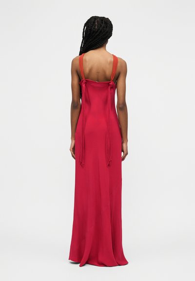 Γυναίκα με πλεξούδες, που φοράει μακρύ κόκκινο φόρεμα μέχρι το πάτωμα με λεπτούς πορτοκαλί ιμάντες και μακριά φουντάκια δεμένα στην πλάτη, στέκεται μπροστά σε απλό φόντο.