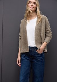 Cardigan beige et duveteux sur un t-shirt blanc, associé à un jean taille haute bleu marine. Le mannequin se tient les mains dans les poches. Fond gris.
