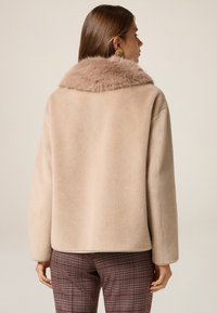 Chaquetón de pelo sintético beige con un cuello acanalado, que presenta una textura suave y una silueta relajada. Combinado con pantalones de cuadros, mostrando colores complementarios.