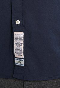 Mörkblå skjortslang med två vita tygmärken; det övre märket har detaljerad varumärkestext, det nedre märket har LA MARTINA-logotypen och texten Buenos Aires.