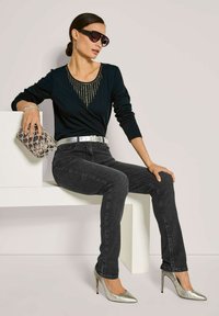 Schwarzes Langarmoberteil mit Detail am Ausschnitt, dunkelgraue Jeans, silberner Gürtel, metallische hohe Absätze und eine strukturierte silberne Clutch.
