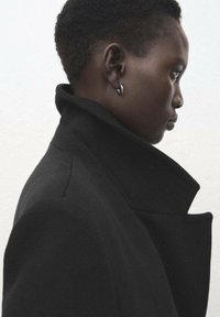 Czarny wełniany płaszcz z wysokim kołnierzem i ostrymi klapami, o gładkiej strukturze. Widoczne ucho z kolczykiem-kołem.