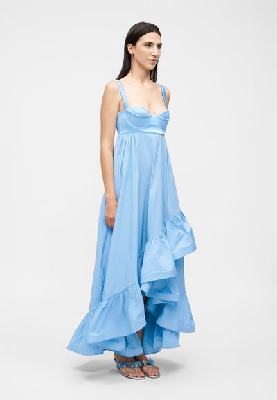 Γυναίκα που φοράει ένα ανοιχτό μπλε, ασύμμετρο, αμάνικο φόρεμα με βολάν στο τελείωμα και σανδάλια, στέκεται μπροστά σε ένα μονόχρωμο λευκό φόντο.